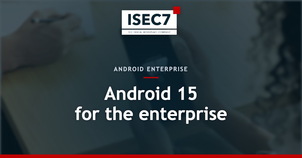 A person holds a smartphone displaying the Android 15 logo on the screen. In the background, a second person is seen writing on a notepad with a pen. The scene takes place at a table in an office setting.